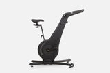 Side view of a black wooden indoor cycle with a black saddle, mesh flywheel cover, and a handlebar touchscreen